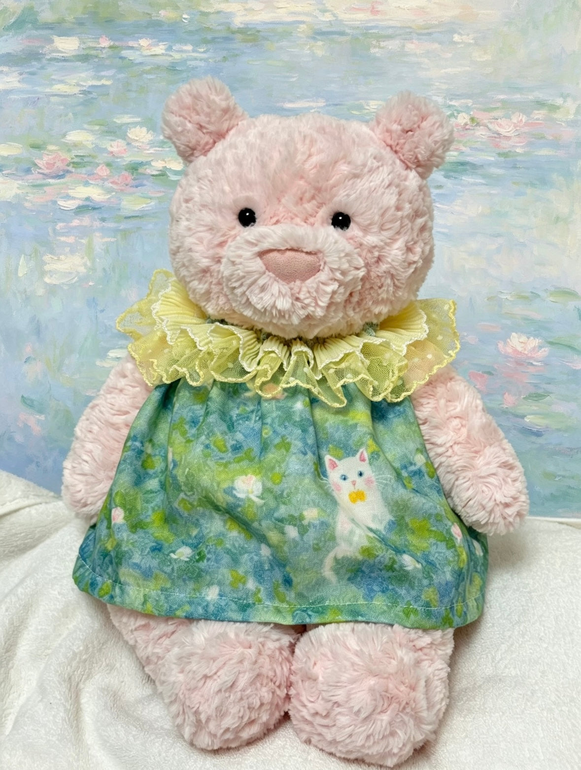 Pale plush bear wearing a blue-and-green watercolour kitten-print dress with a pale yellow-green ruffled lace collar
