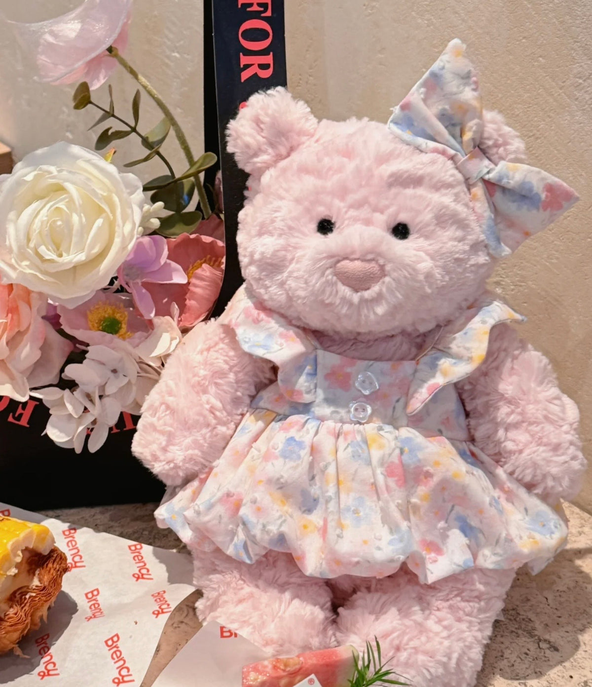 Pink plush bear modelling a pastel floral dress and matching ear bow beside flowers