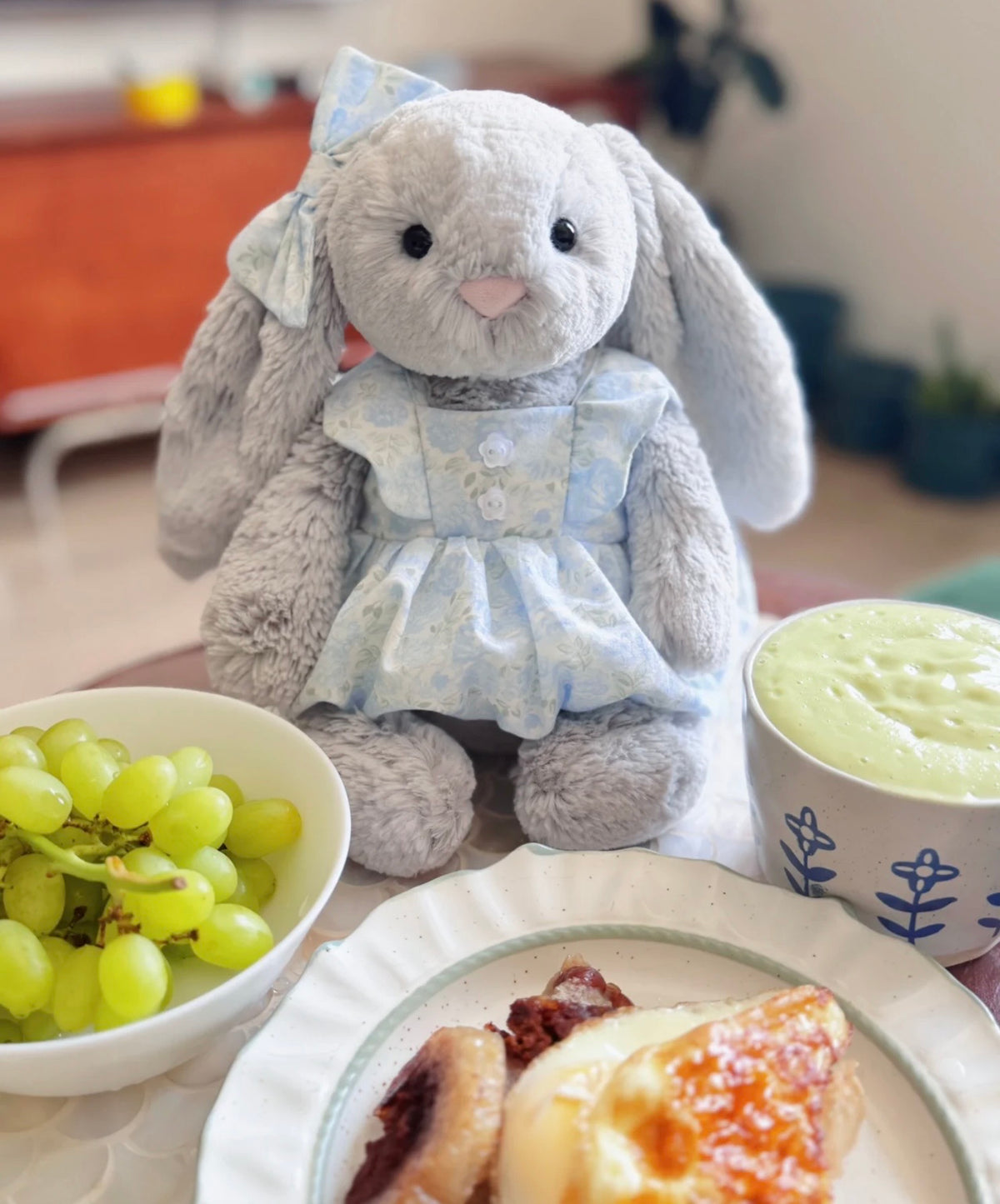 Grey plush bunny wearing a blue floral dress and matching bow at a table with afternoon treats