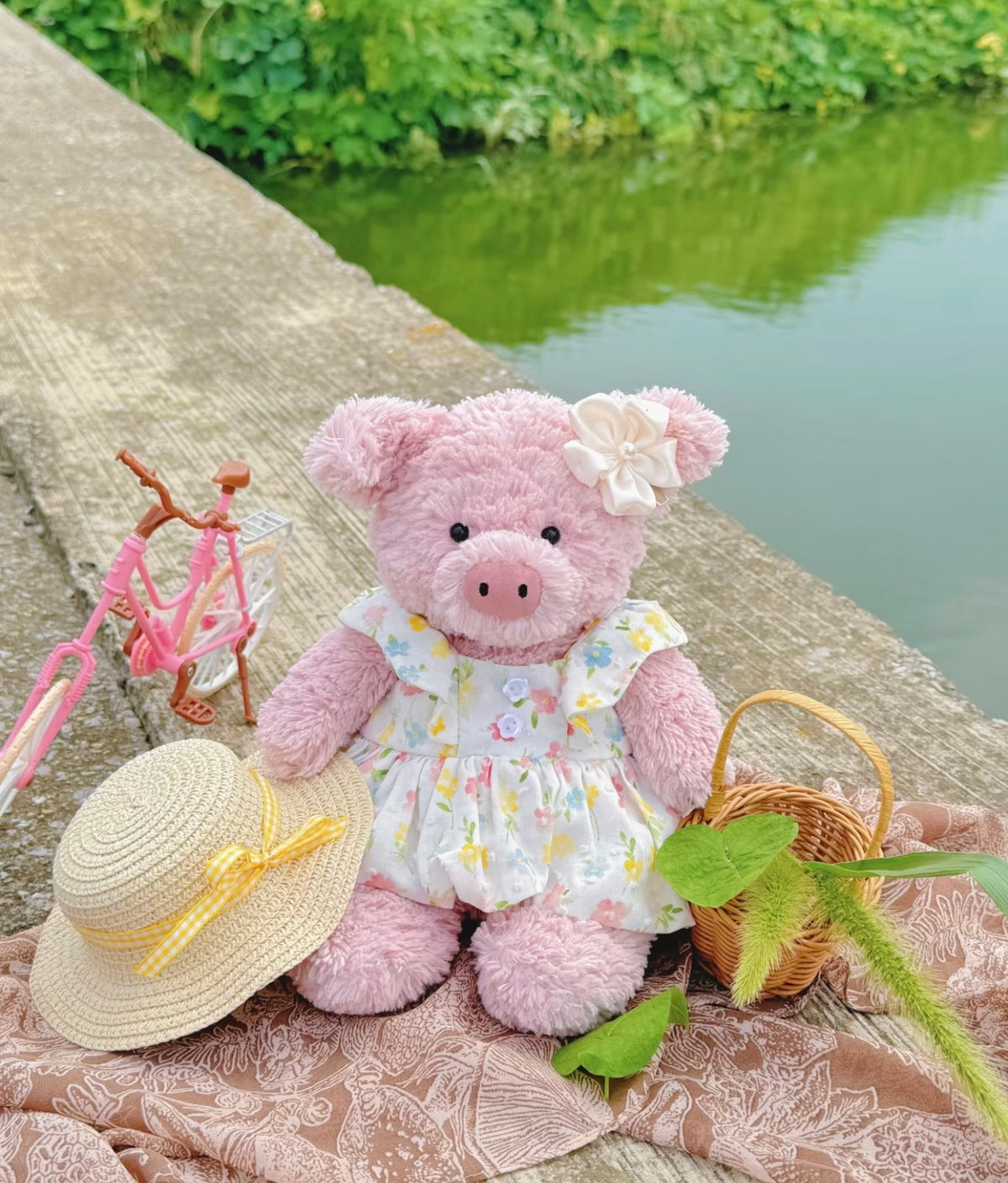Pink plush pig wearing a pastel floral dress and straw-style hat beside a miniature bicycle and basket