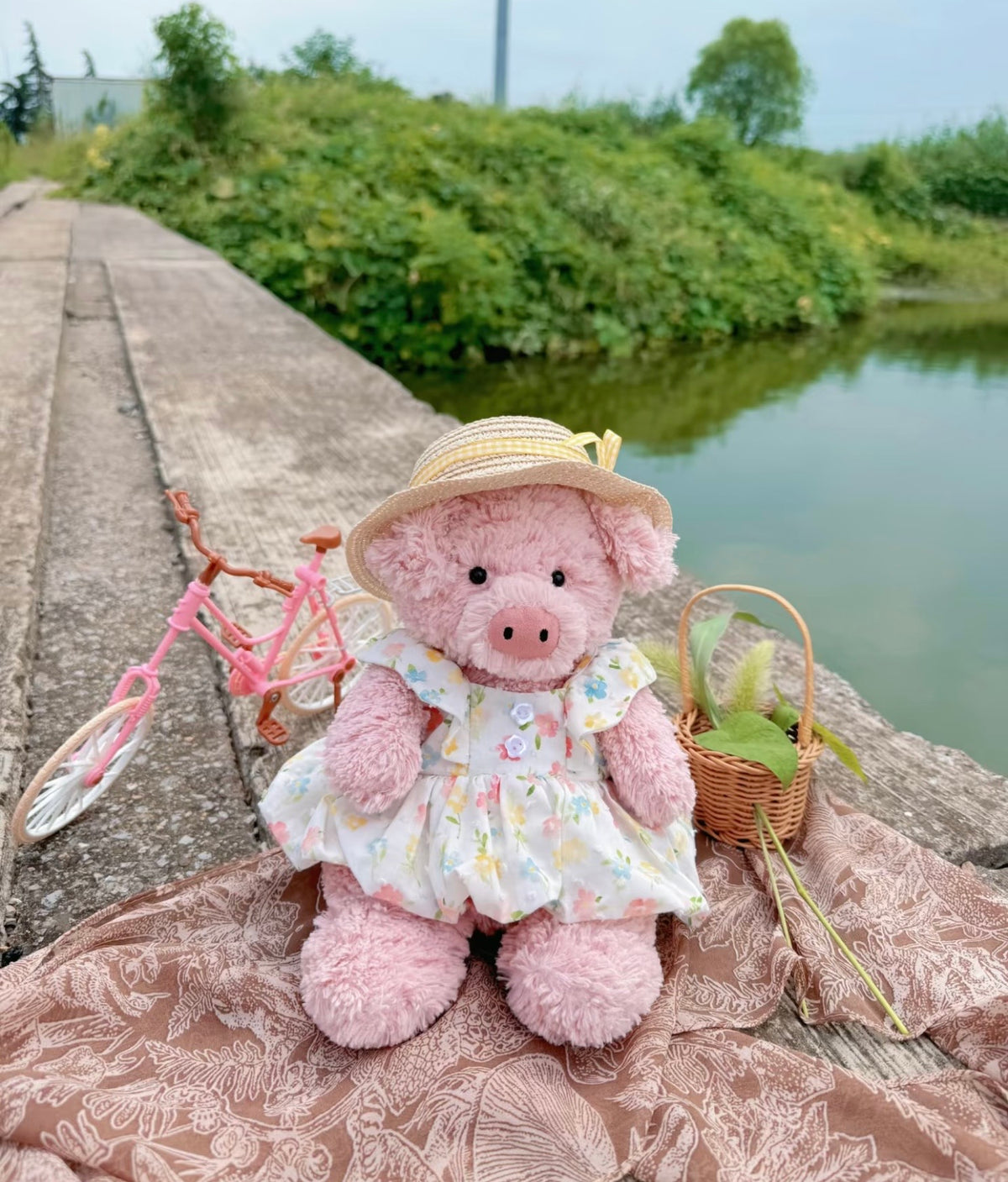 Pink plush pig modelling a pastel floral dress beside a hat and basket in an outdoor picnic scene