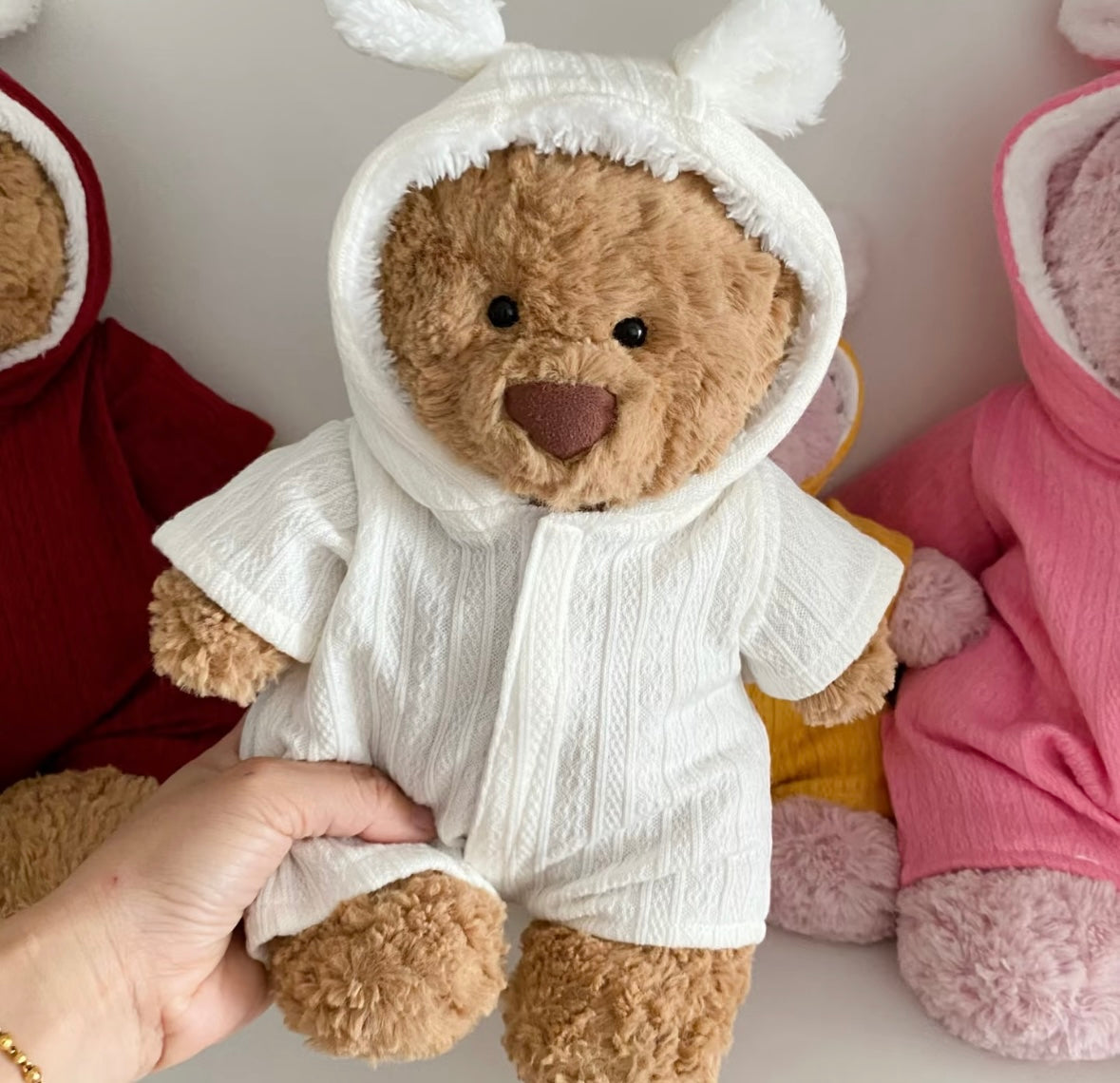 Five plush toys wearing bunny-ear hooded rompers in grey, yellow, cream, blue and pink