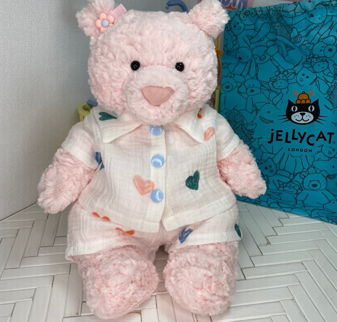 Pink teddy bear wearing cream muslin pyjamas with a multicolour heart print and blue buttons