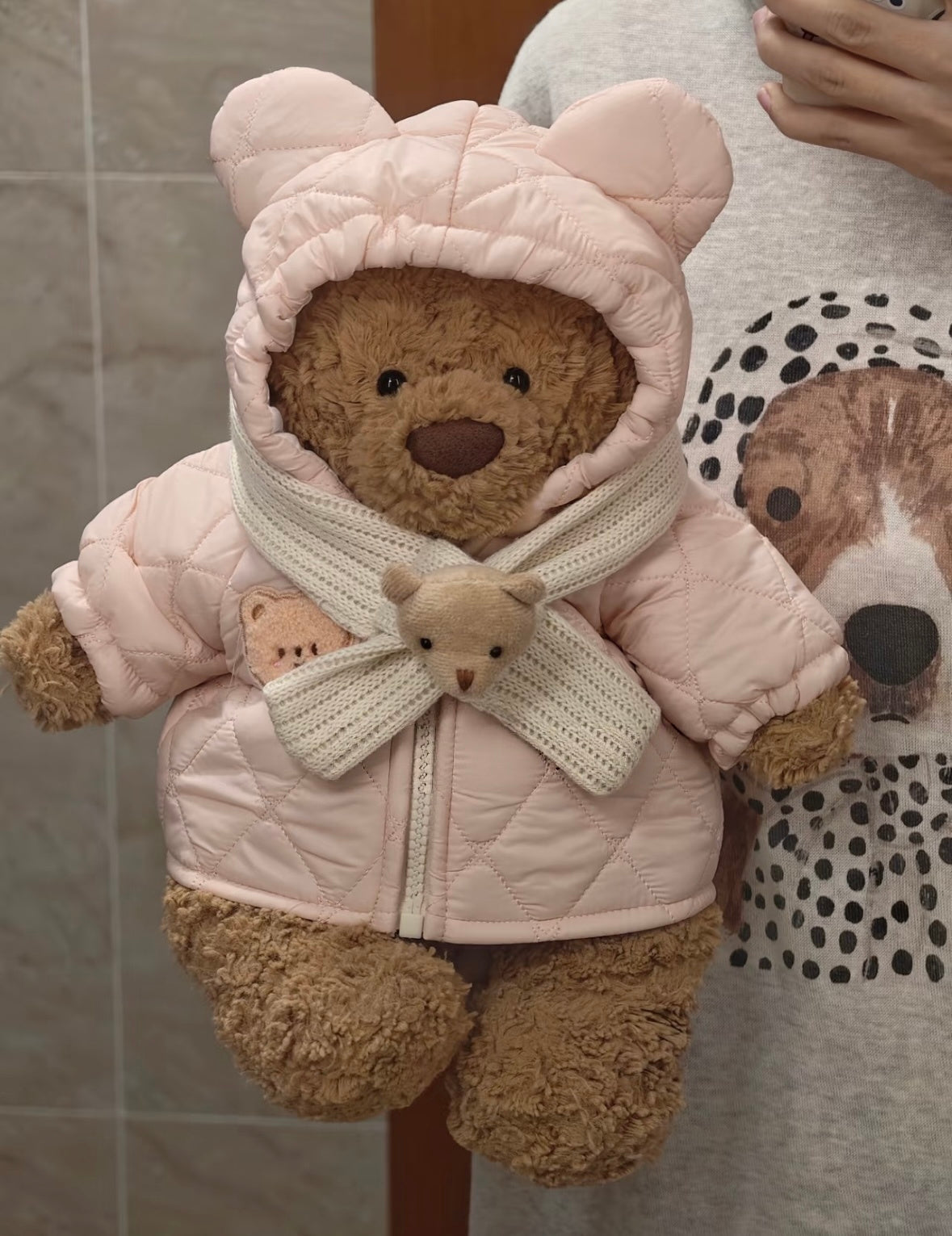 Brown teddy bear wearing a pink quilted hooded coat with bear ears and a knitted scarf