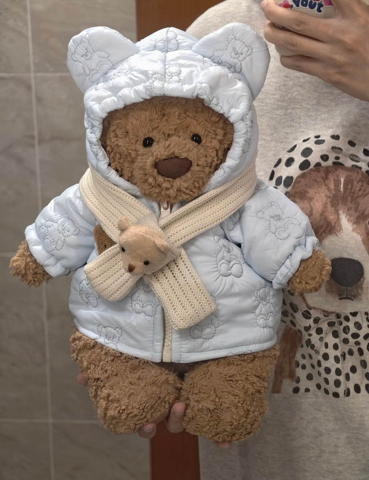 Brown teddy bear wearing a white quilted hooded coat with a bear-ear hood and scarf
