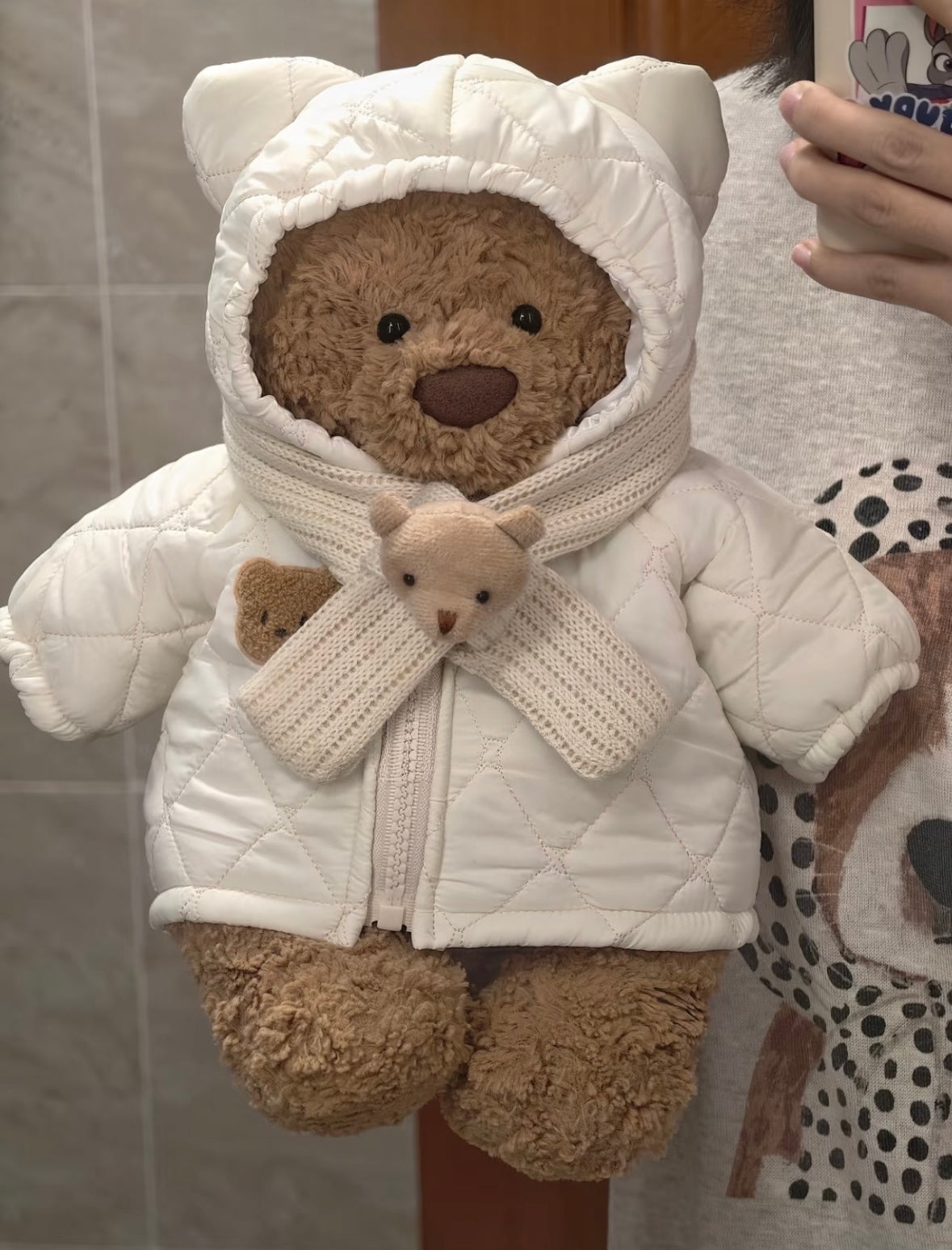 Brown teddy bear wearing a pale blue quilted hooded coat with a bear-ear hood and scarf