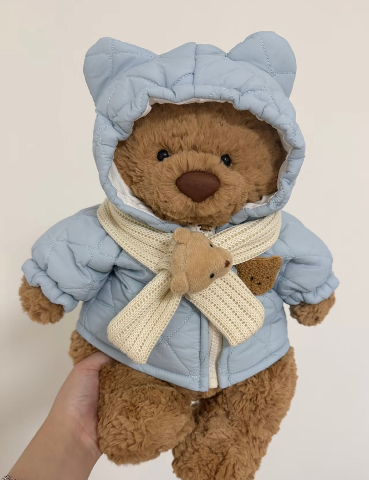 Brown teddy bear wearing a cream quilted hooded coat with a teddy bear scarf detail