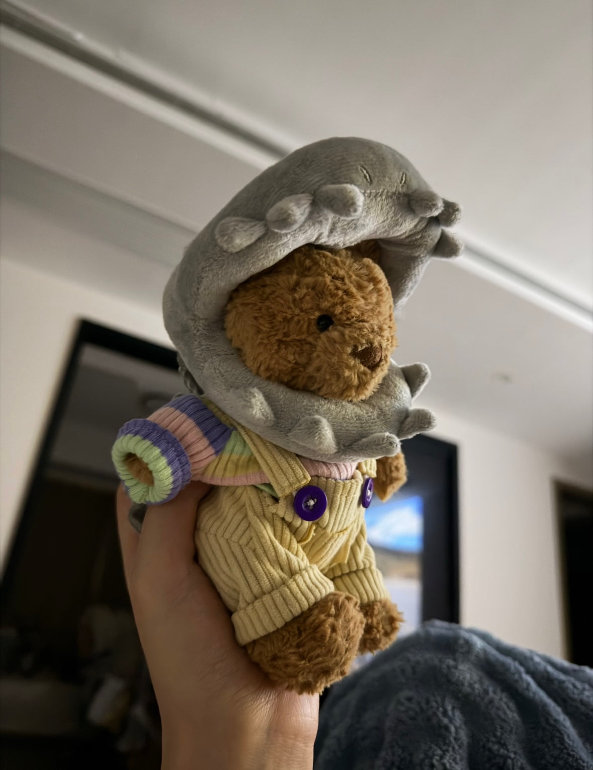 Bartholomew Bear wearing a grey shark headpiece, pale yellow dungarees with purple buttons and a pastel striped top