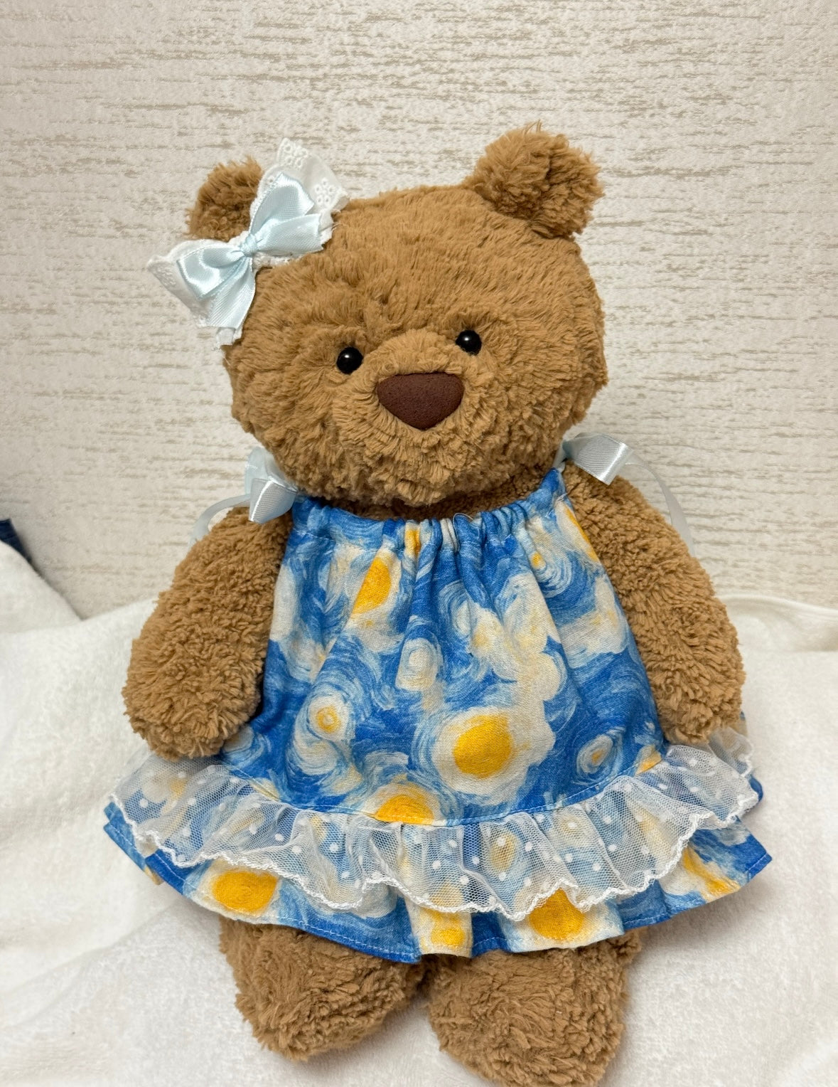 Close view of Bartholomew Bear's blue printed dress, pale shoulder ribbons and dotted lace hem