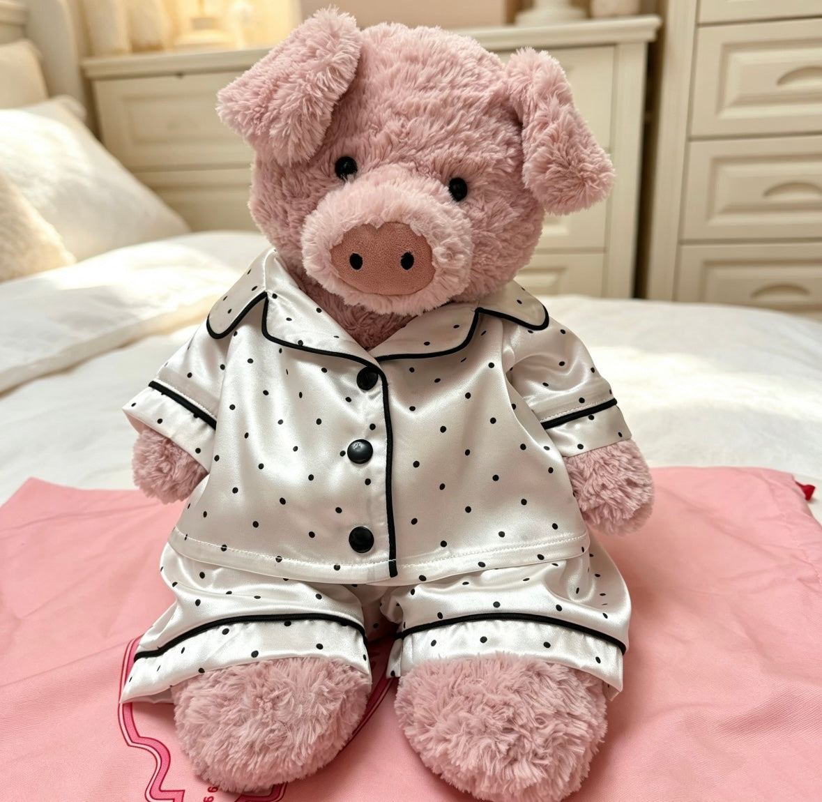 Pink plush pig seated on a bed wearing the matching ivory and black polka-dot pyjama set