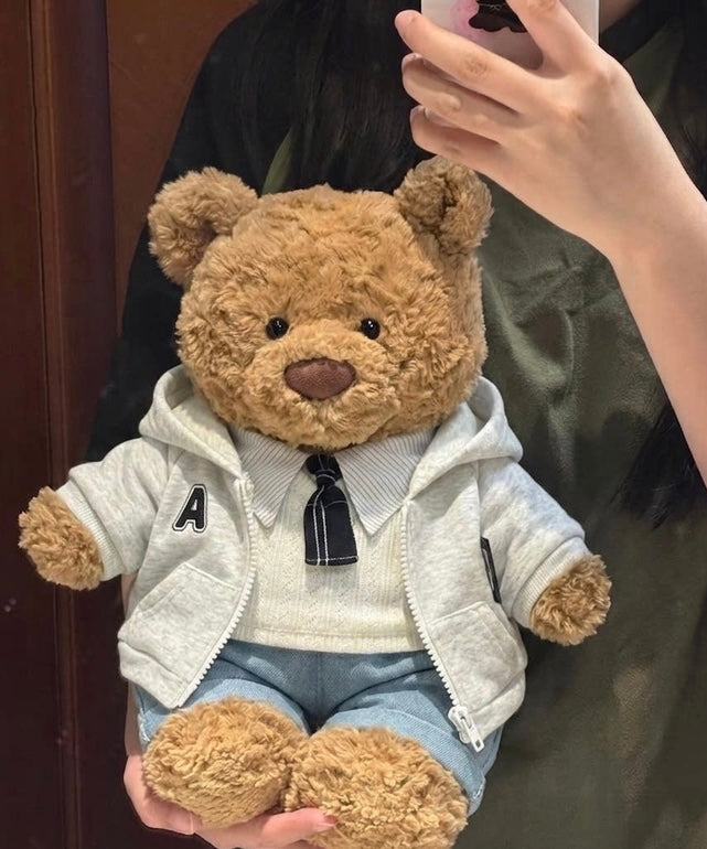 Bartholomew Bear wearing a light grey A-patch hoodie, cream top with striped collar and dark tie, and light blue bottoms