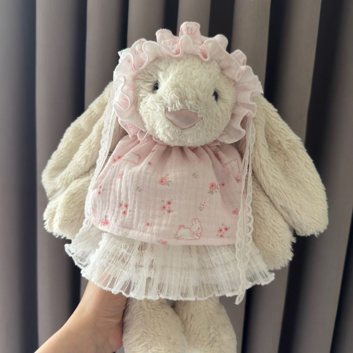 Cream Bashful Bunny wearing a pale pink printed dress with cream lace skirt and a matching ruffled bonnet with lace ties