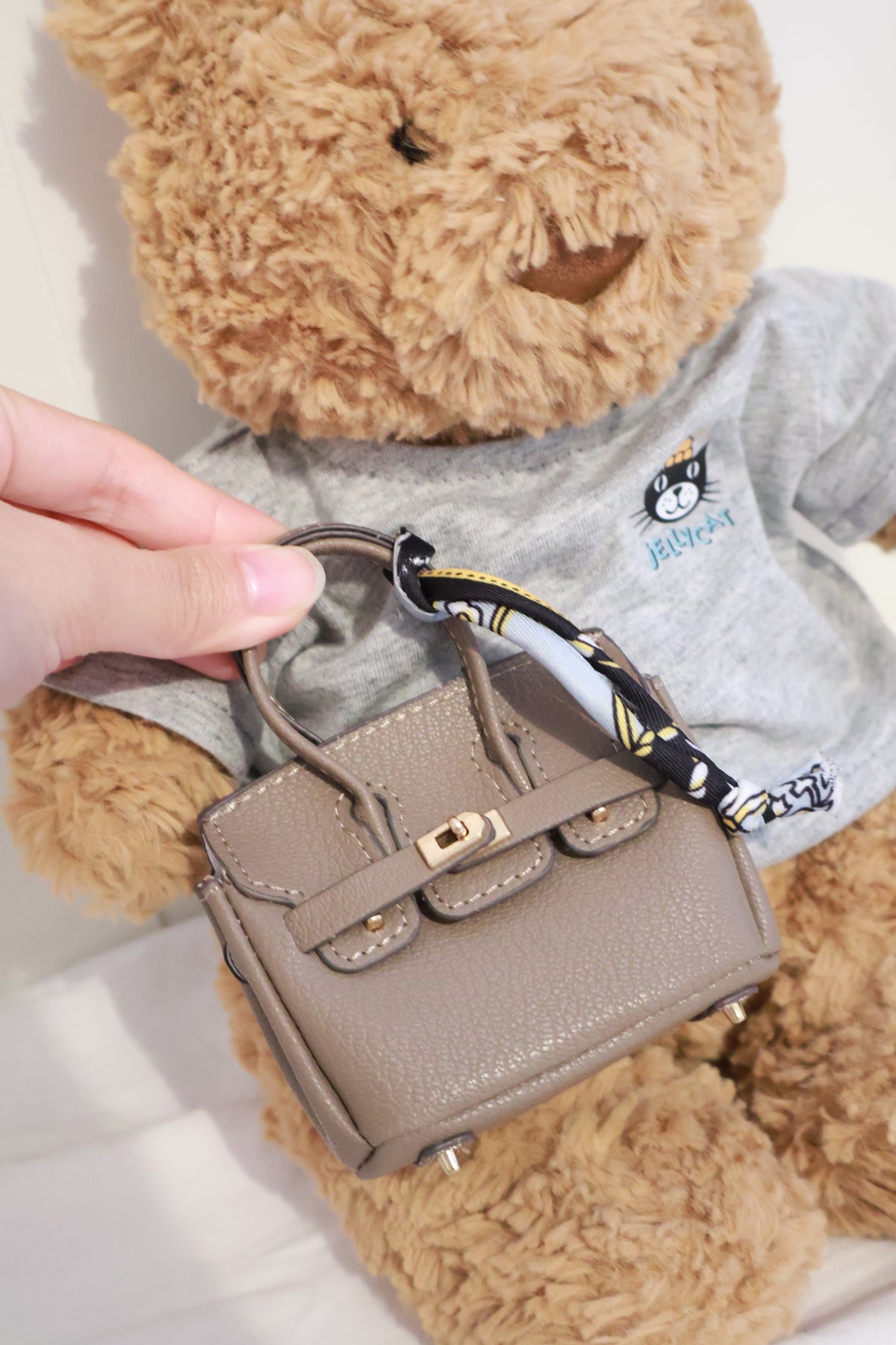 Taupe miniature handbag with twin handles, gold-tone clasp and patterned handle scarf displayed in front of a plush bear wearing a grey top