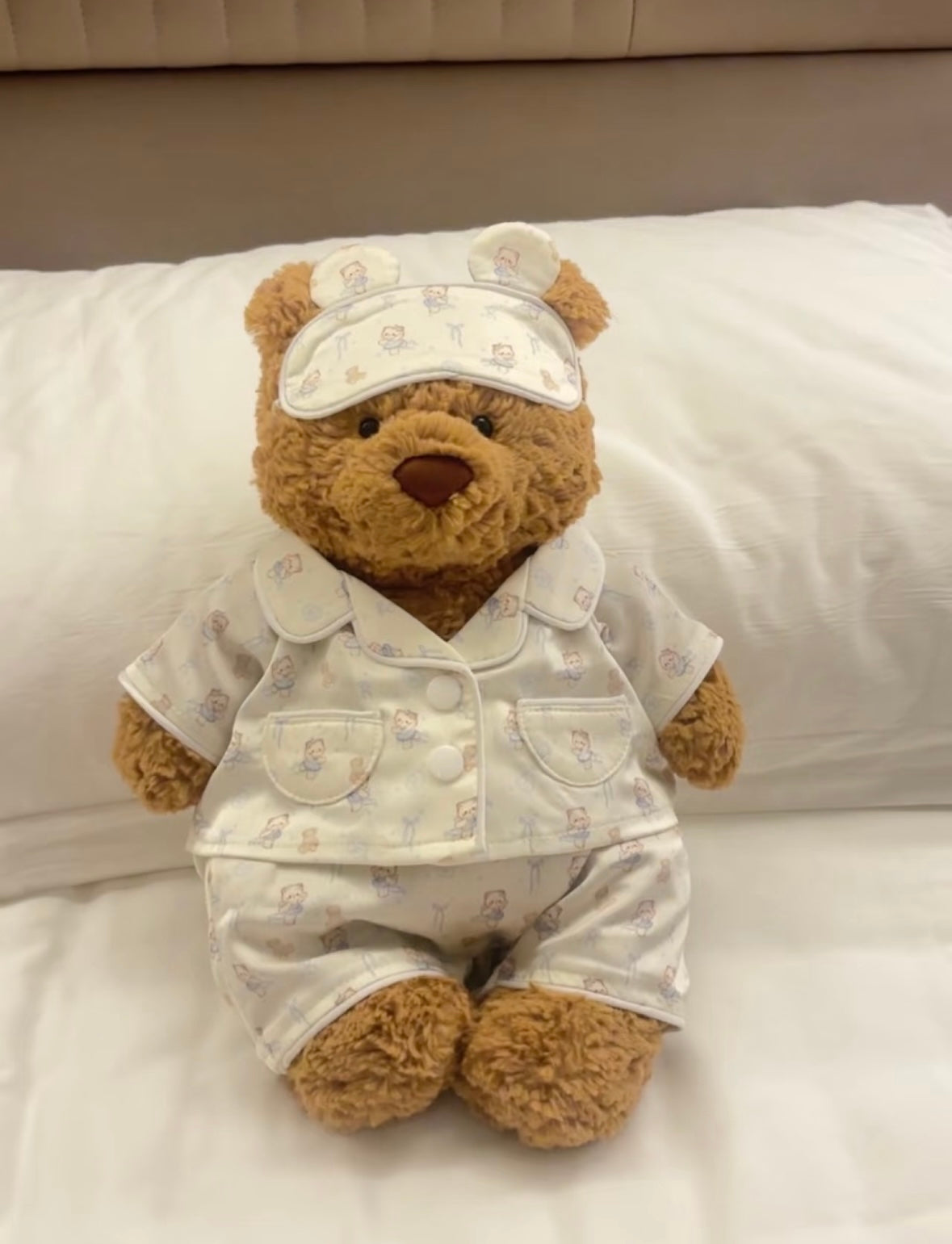Bartholomew Bear wearing pyjama set and sleep mask, sitting upright on white bedding
