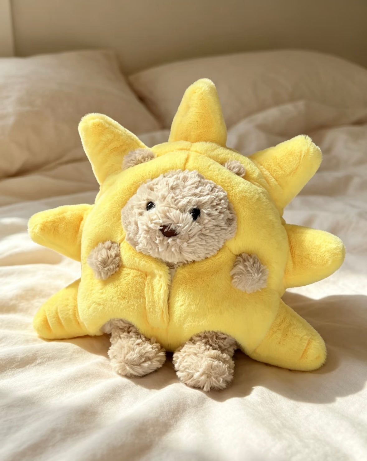 Bartholomew Bear wearing a pale yellow plush sunshine costume with padded sun rays, seated on white bedding
