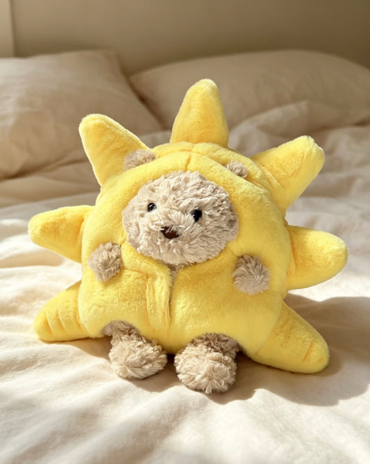 Bartholomew Bear wearing a pale yellow plush sunshine costume with padded sun rays, seated on white bedding