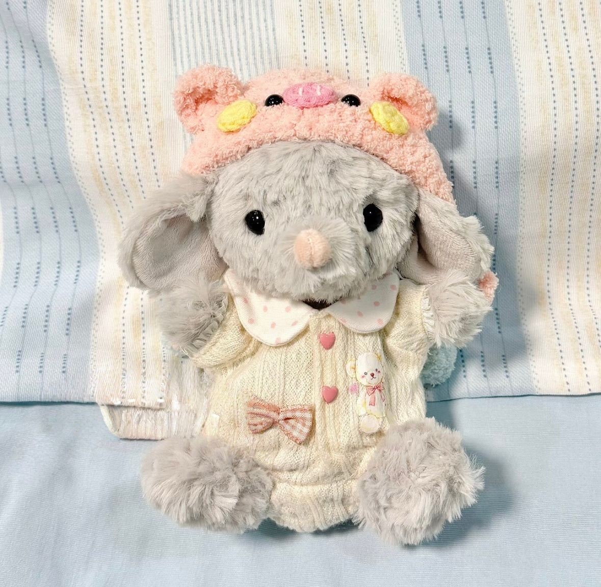 Grey elephant plush wearing a cream knit dress with peter pan collar, bunny embroidery and fluffy pink bear hat with yellow flowers