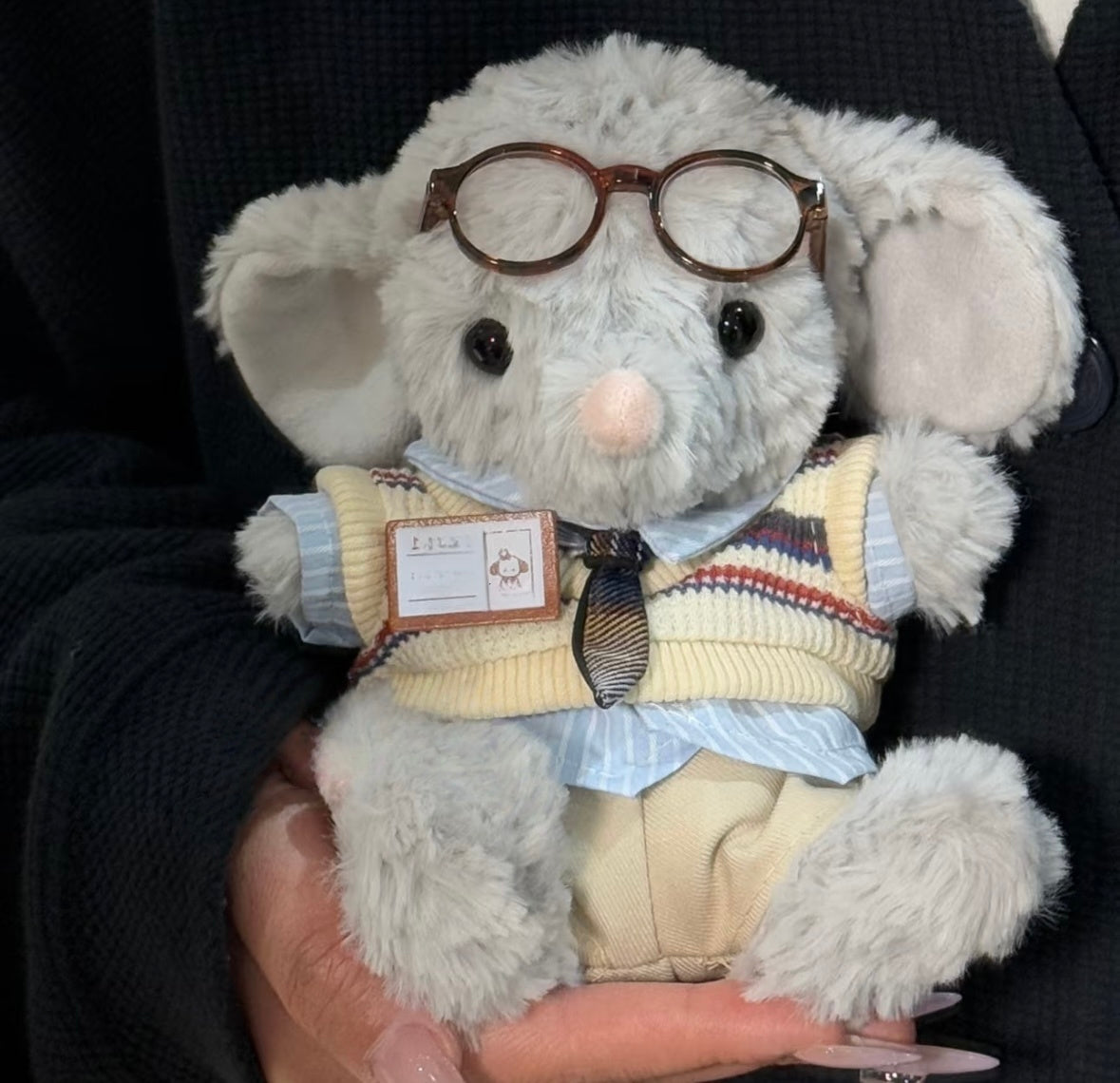 Grey elephant plush wearing round tortoiseshell glasses, cream varsity sweater vest over blue striped shirt with plaid tie and name badge