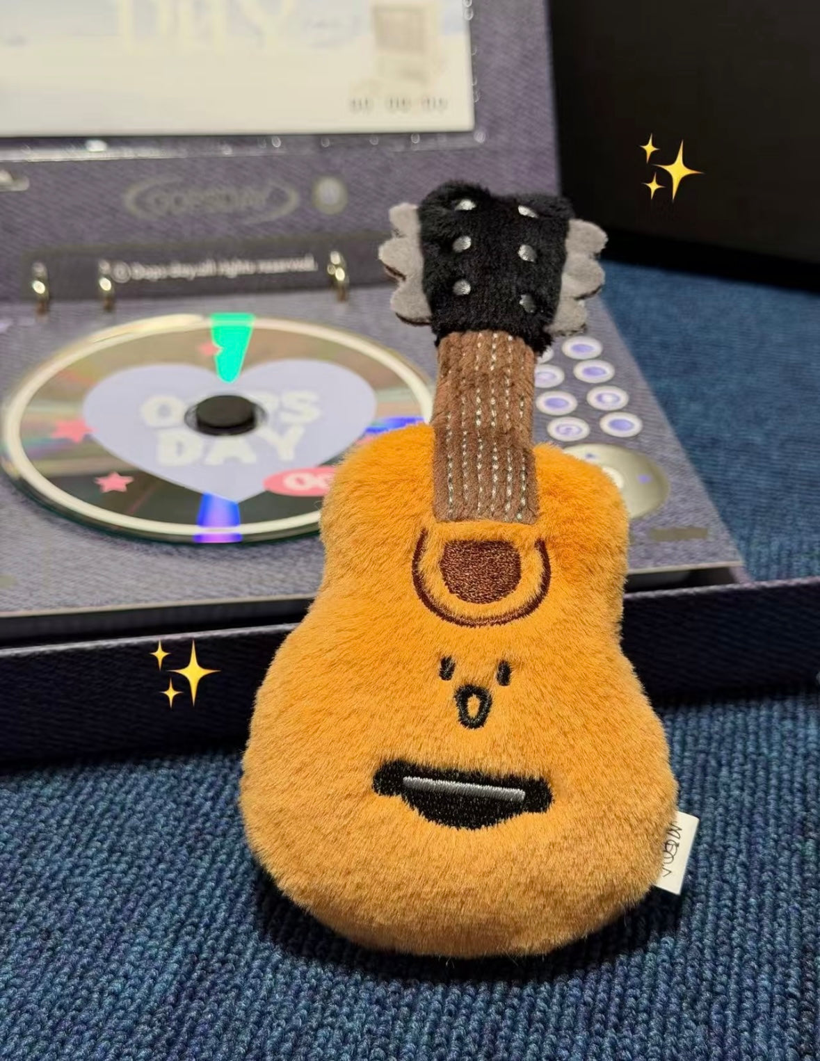 Fluffy golden guitar plush keychain with embroidered face propped against a CD player