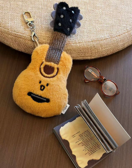 Fluffy golden guitar plush keychain with silver clasp laid flat next to glasses and a book
