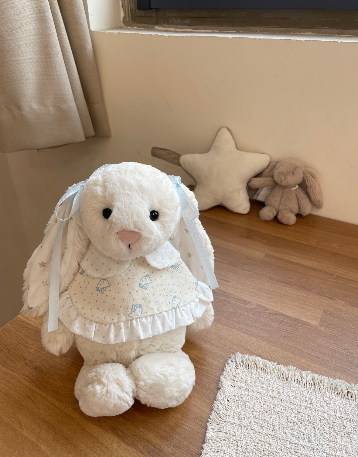 White Bashful Bunny wearing blue bunny print dress with ribbon bows - seated on wooden surface