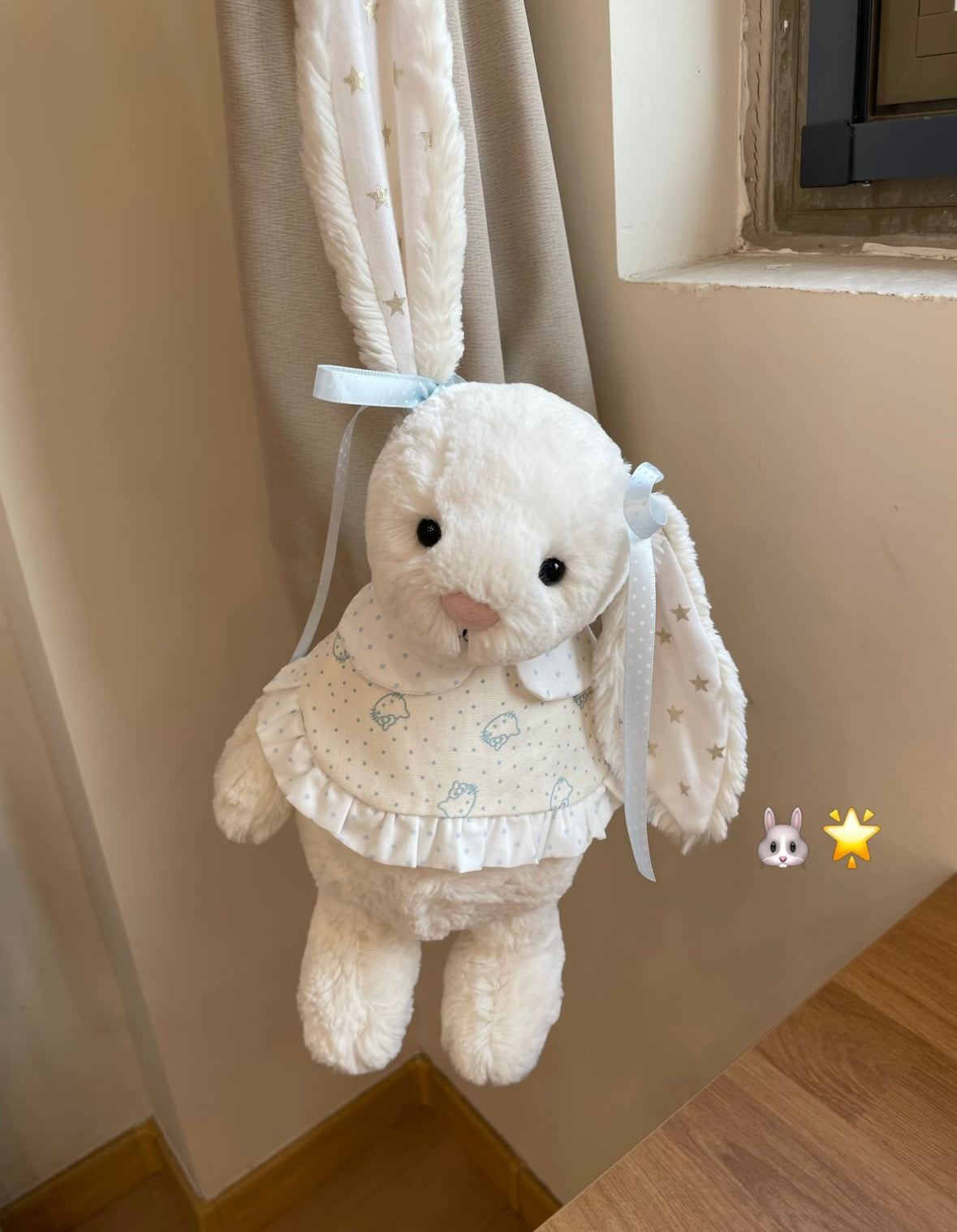 White Bashful Bunny wearing blue bunny print dress - hanging against curtain, front view