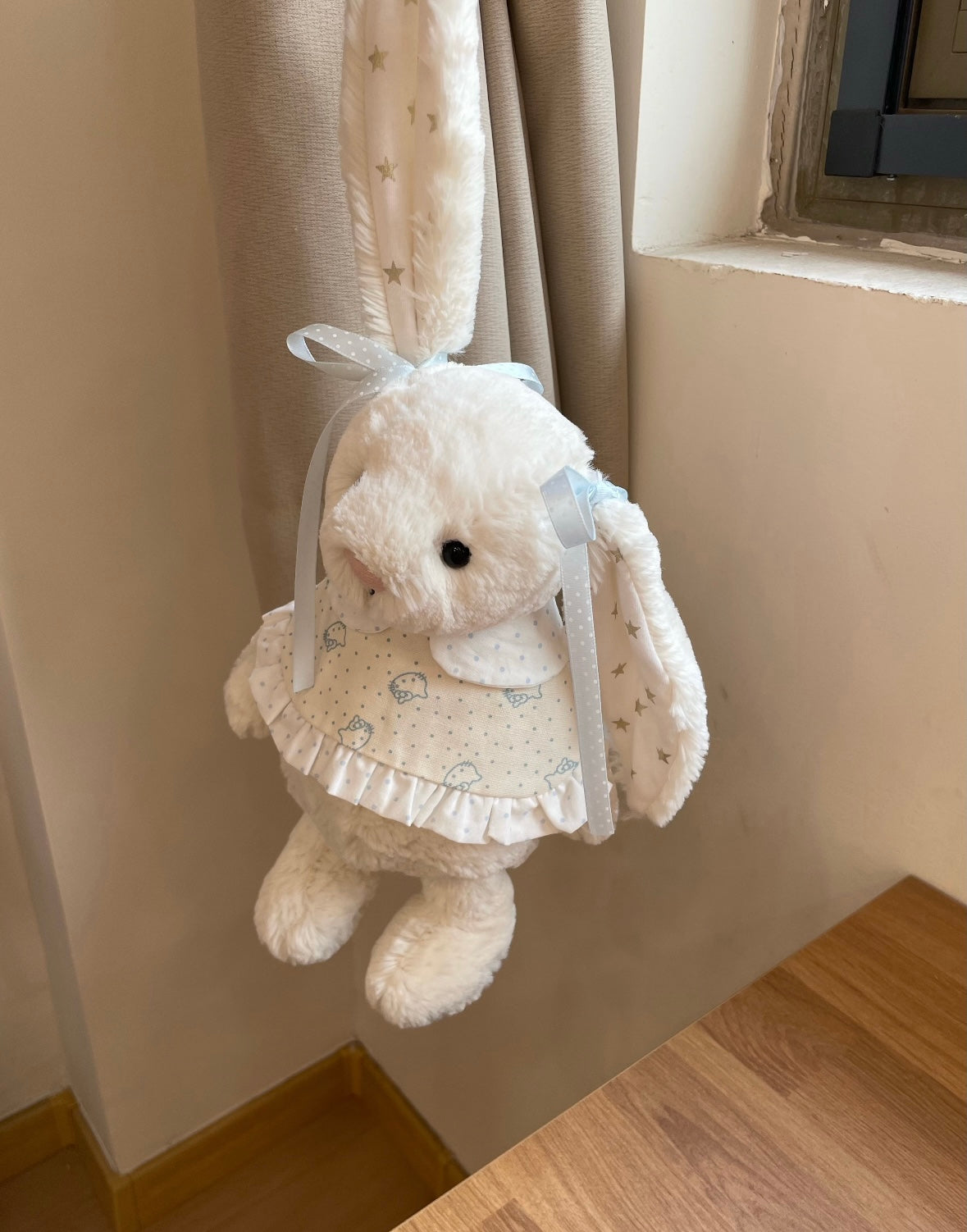 White Bashful Bunny wearing blue bunny print dress - hanging against curtain, side view