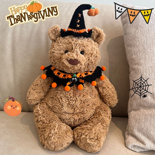 Plush bear wearing a black crochet witch hat with mini pumpkin charm and an orange pompom collar with spider centrepiece