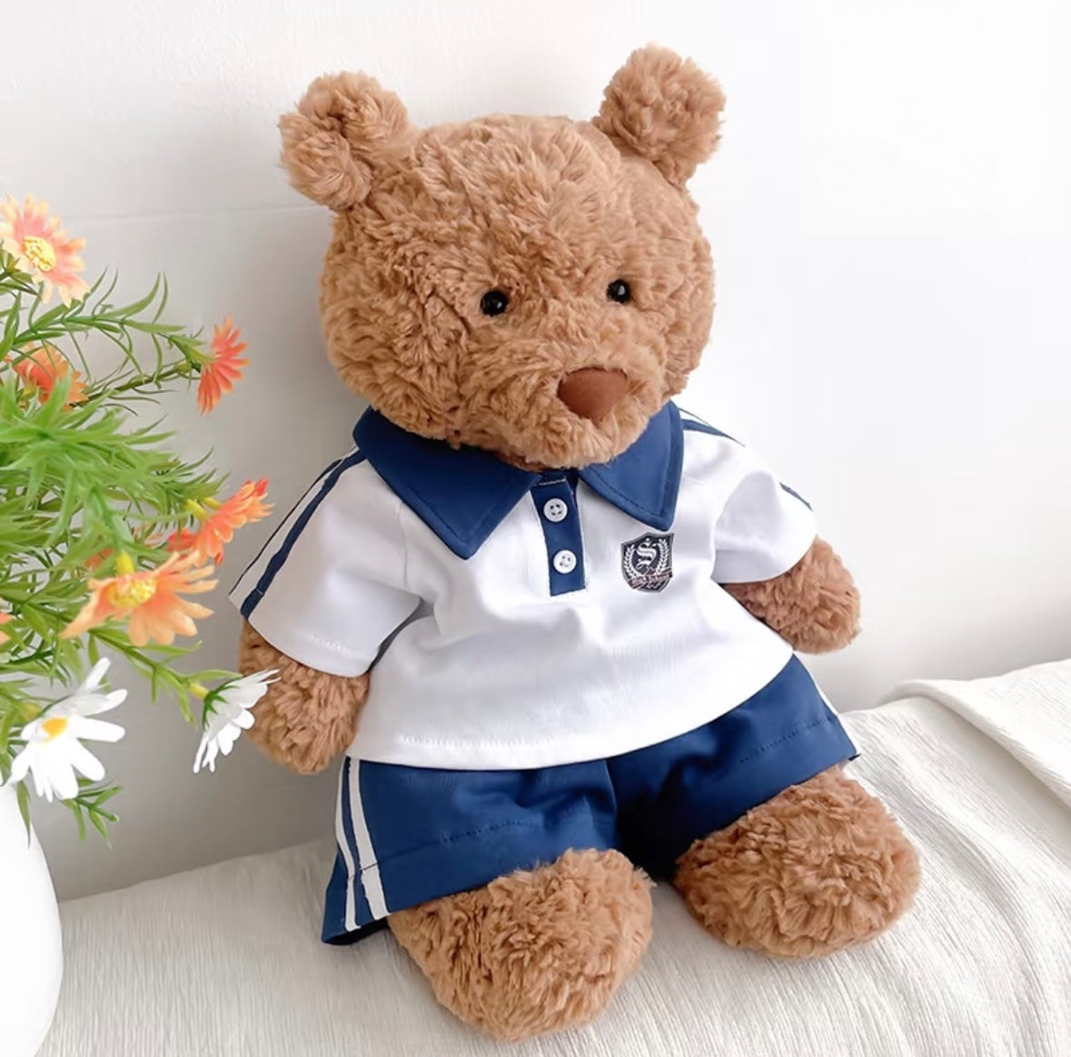 Brown bear plush in navy polo sport set seated against white drape with flowers