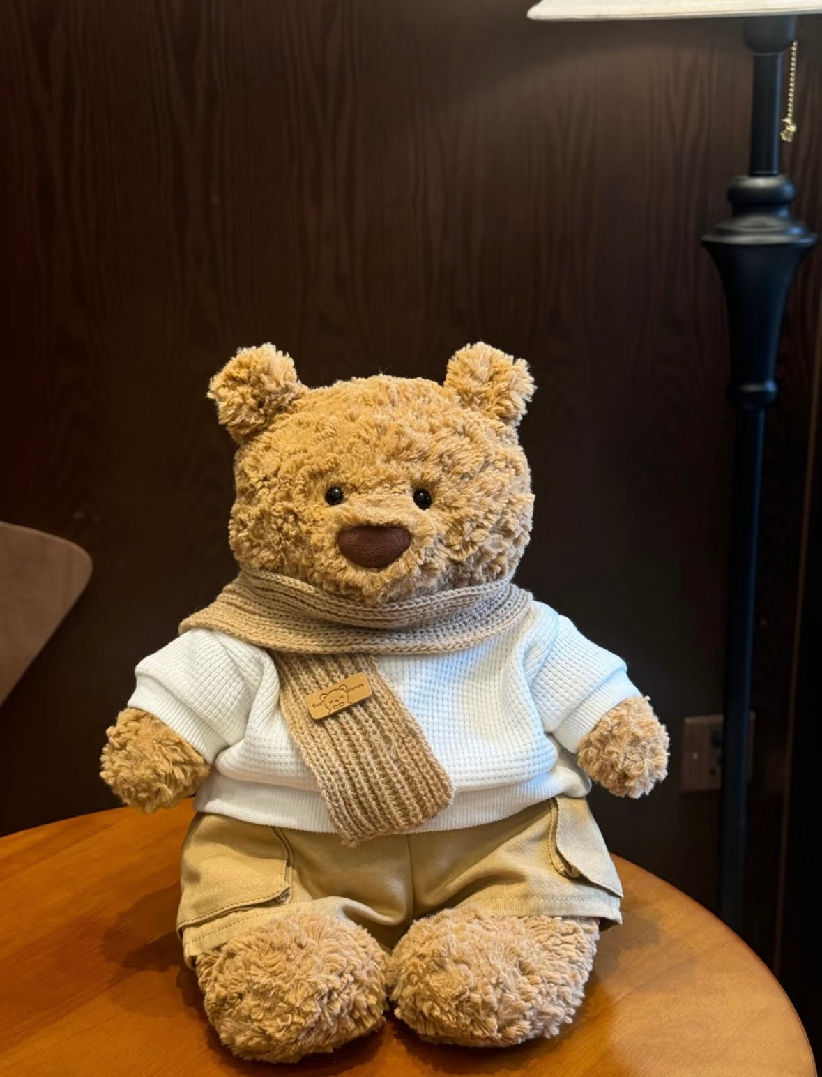 Brown bear plush wearing white waffle-knit sweater, oatmeal ribbed scarf and khaki shorts, seated on wooden table