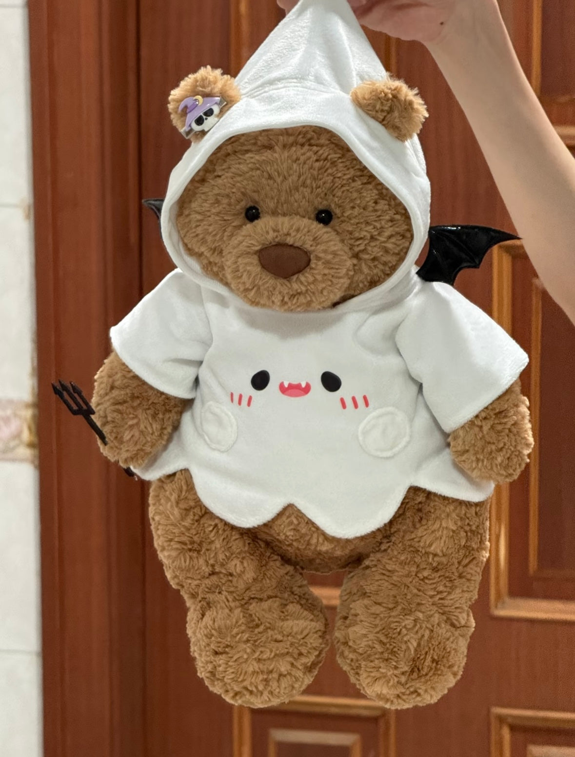 Brown plush bear wearing white ghost hoodie with embroidered vampire face, black bat wings and mini devil trident