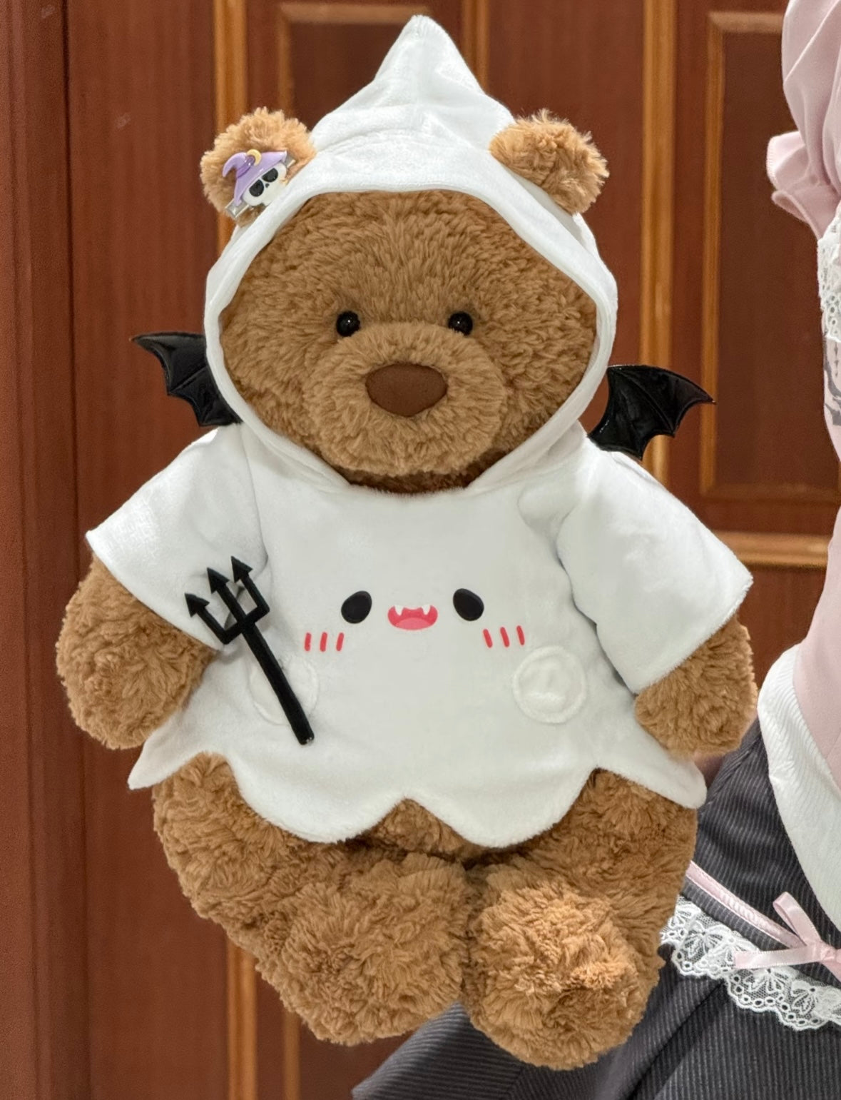 Full view of plush bear wearing white ghost hoodie Halloween costume with bat wings and devil trident prop