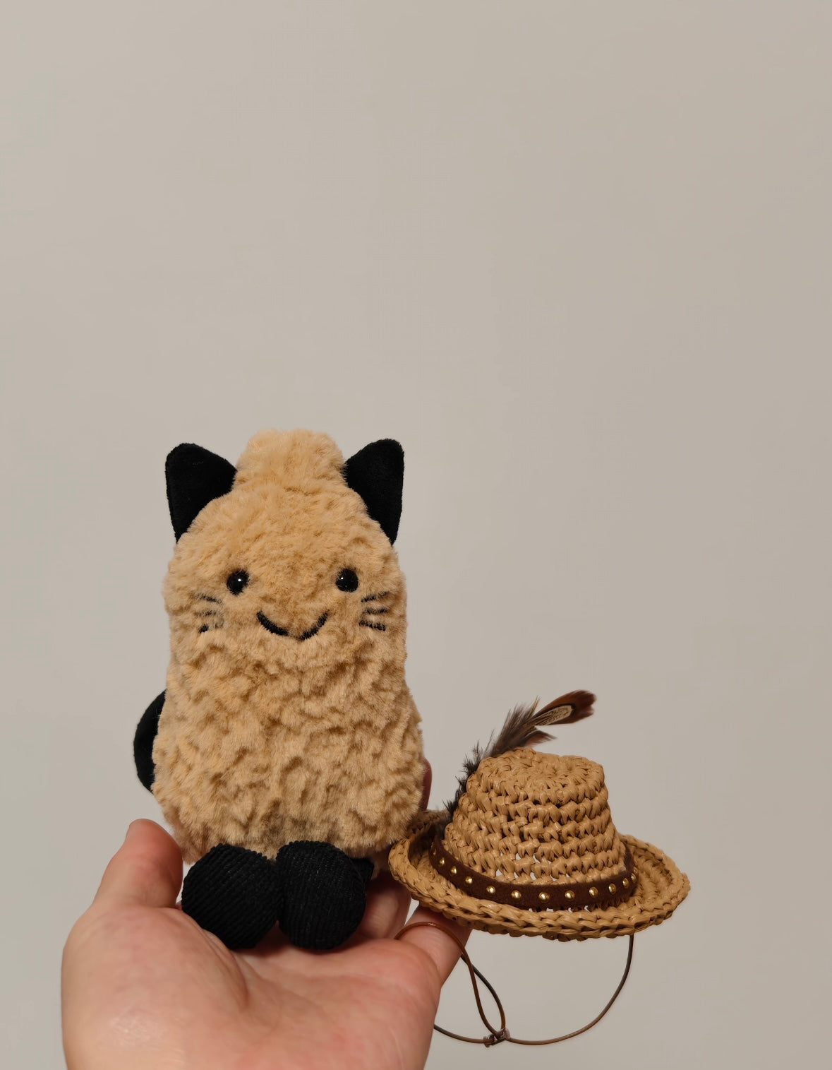 Jellycat Amuseable cat plush holding a mini woven straw cowboy hat with feather and gold bead trim