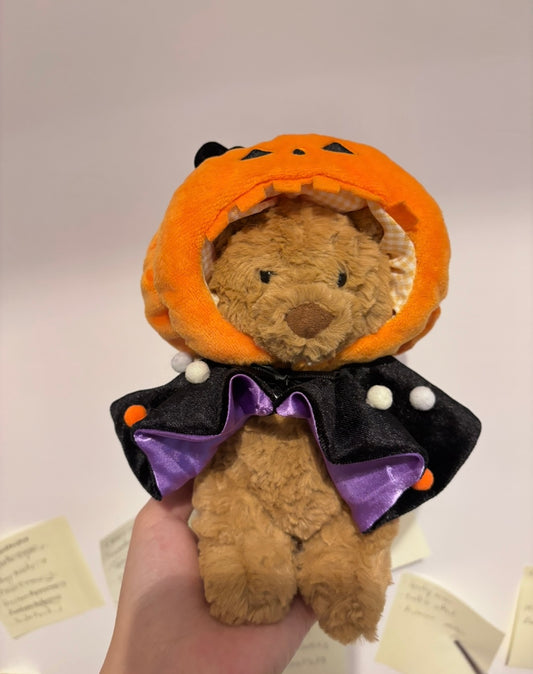 Brown plush bear wearing orange pumpkin hat and black cape with purple bow - front view