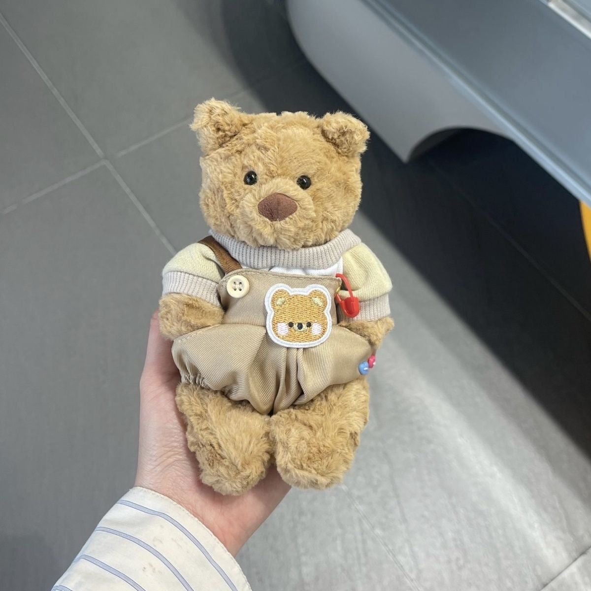 Brown plush bear wearing khaki dungarees with cream turtleneck jumper, bear patch and charm accessories
