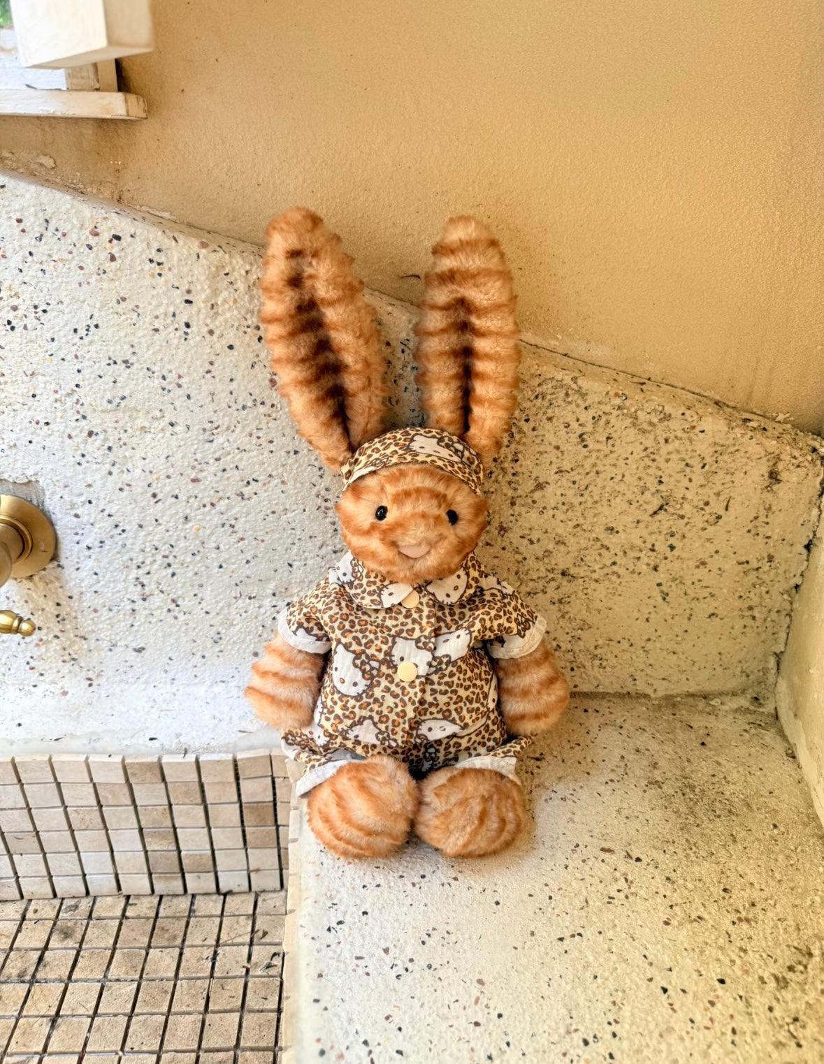 Jellycat bunny in Hello Kitty leopard print outfit seated outdoors on ledge