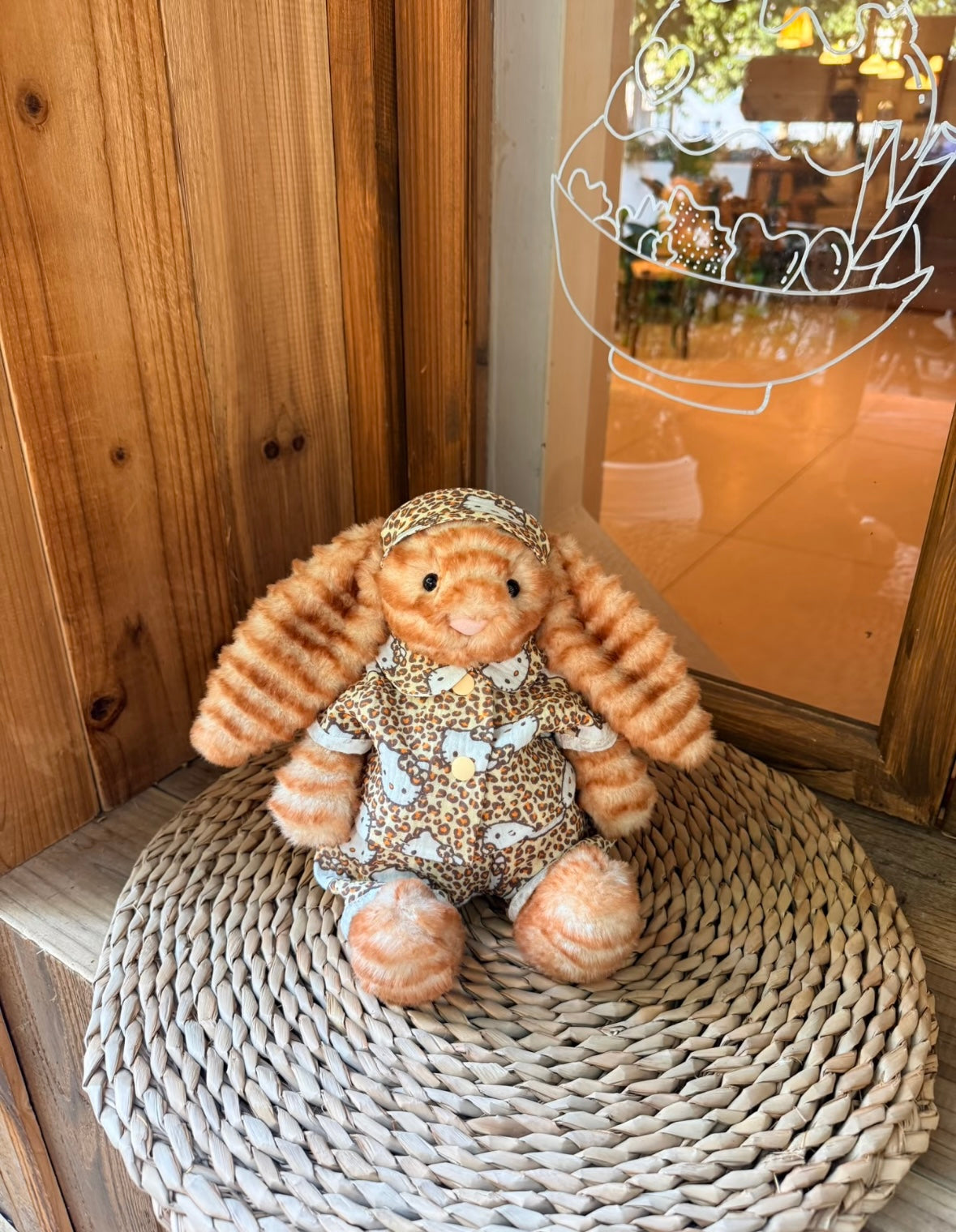 Jellycat bunny wearing Hello Kitty leopard print set, seated on wicker mat by shop window