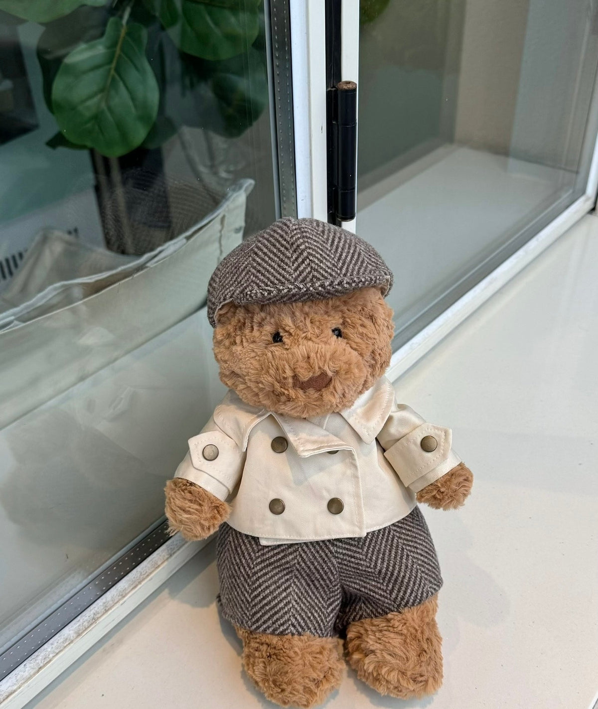 Bartholomew Bear wearing cream trench coat, grey herringbone trousers and flat cap, seated by window